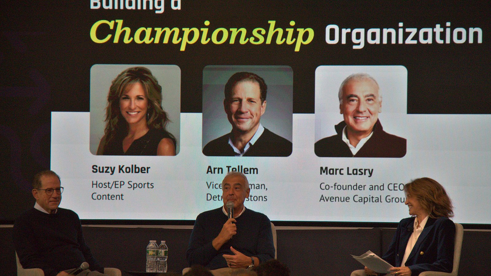 Marc Lasry speaks into a microphone between Arn Tellem and Suzy Kolber under a Building a Championship Organization slide